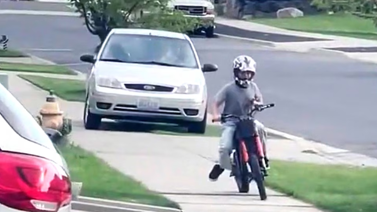 A person wearing a helmet rides a red motorcycle along a residential sidewalk, performing a wheelie with the front wheel lifted off the ground, while a white car is parked on the street nearby.