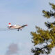 Small crop-duster airplane flies past a green tree in a blue sky, leaving a vapor trail behind it.