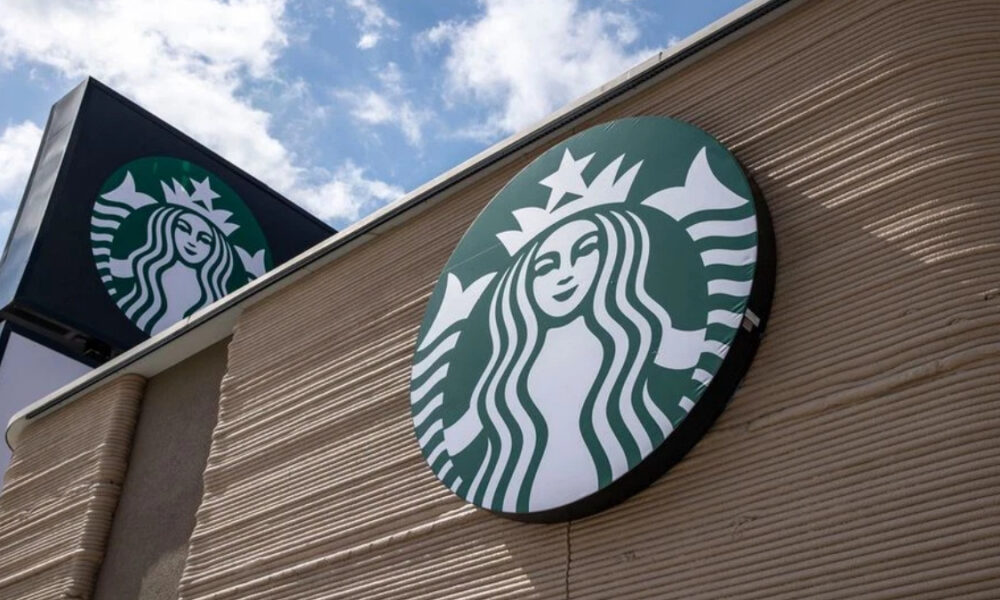 Exterior Starbucks storefront sign featuring the green circular siren logo on a beige brick wall against a blue sky