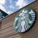 Exterior Starbucks storefront sign featuring the green circular siren logo on a beige brick wall against a blue sky