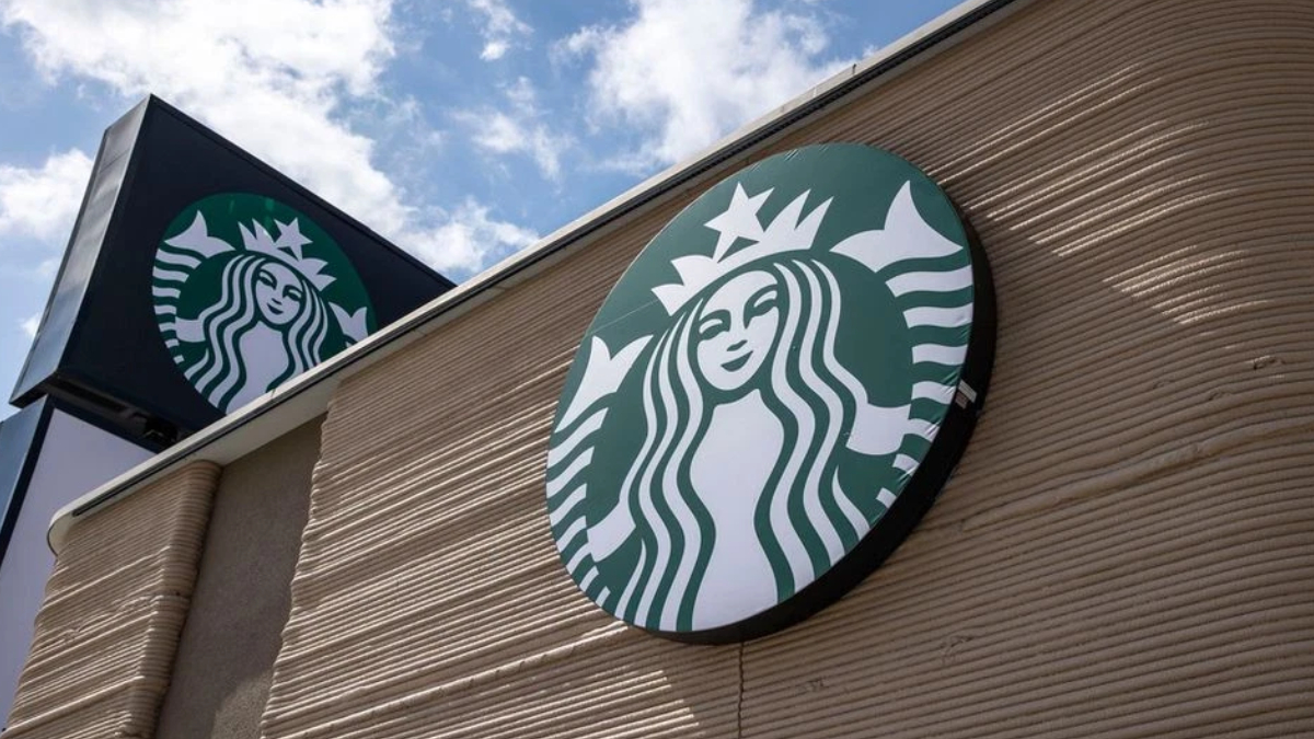 Exterior Starbucks storefront sign featuring the green circular siren logo on a beige brick wall against a blue sky