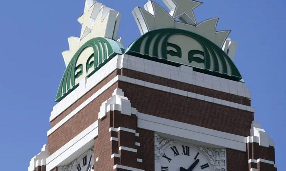 Brick clock tower with a large green circular emblem on top and a Roman numeral clock below.