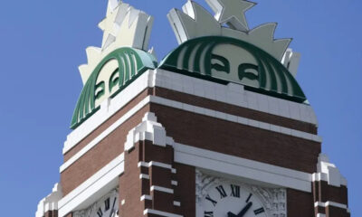 Brick clock tower with a large green circular emblem on top and a Roman numeral clock below.