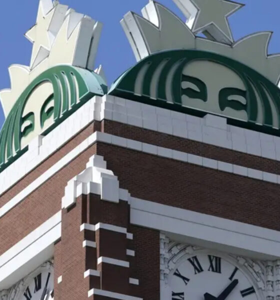 Brick clock tower with a large green circular emblem on top and a Roman numeral clock below.