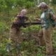Two people in a wooded, overgrown area lift a long black pipe between them, wearing gloves and work clothes.
