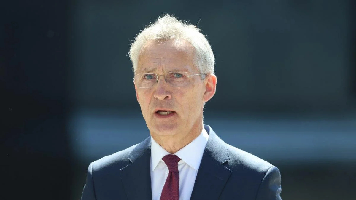 Older man in a dark suit and red tie speaking outdoors