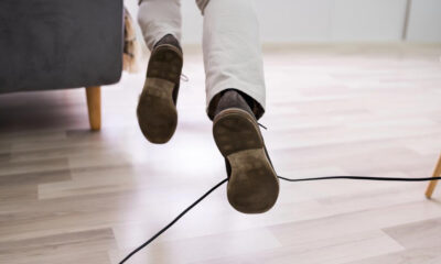 A person walking indoors, seen from behind, with brown shoes and a black cable running across a light wooden floor.