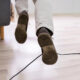 A person walking indoors, seen from behind, with brown shoes and a black cable running across a light wooden floor.