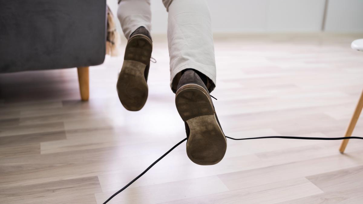 A person walking indoors, seen from behind, with brown shoes and a black cable running across a light wooden floor.