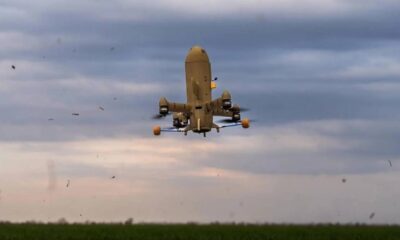 Large quadcopter UAV with orange landing gear hovering low over a grassy field under a cloudy sky