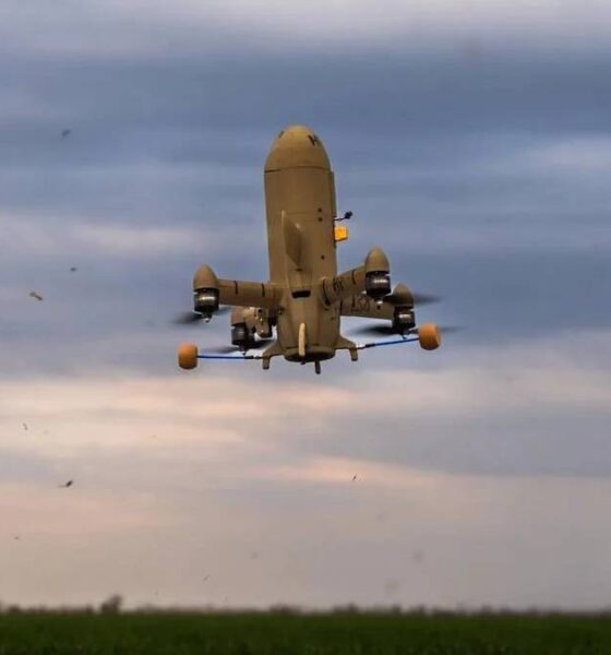 Large quadcopter UAV with orange landing gear hovering low over a grassy field under a cloudy sky