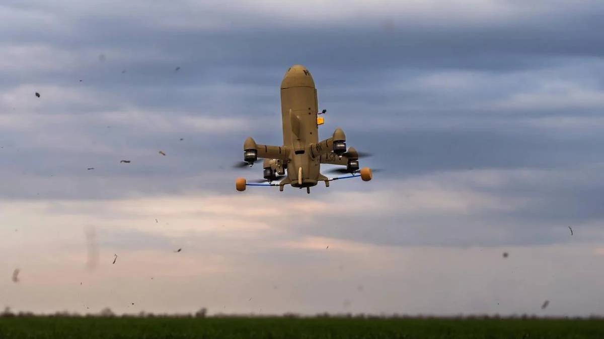 Large quadcopter UAV with orange landing gear hovering low over a grassy field under a cloudy sky