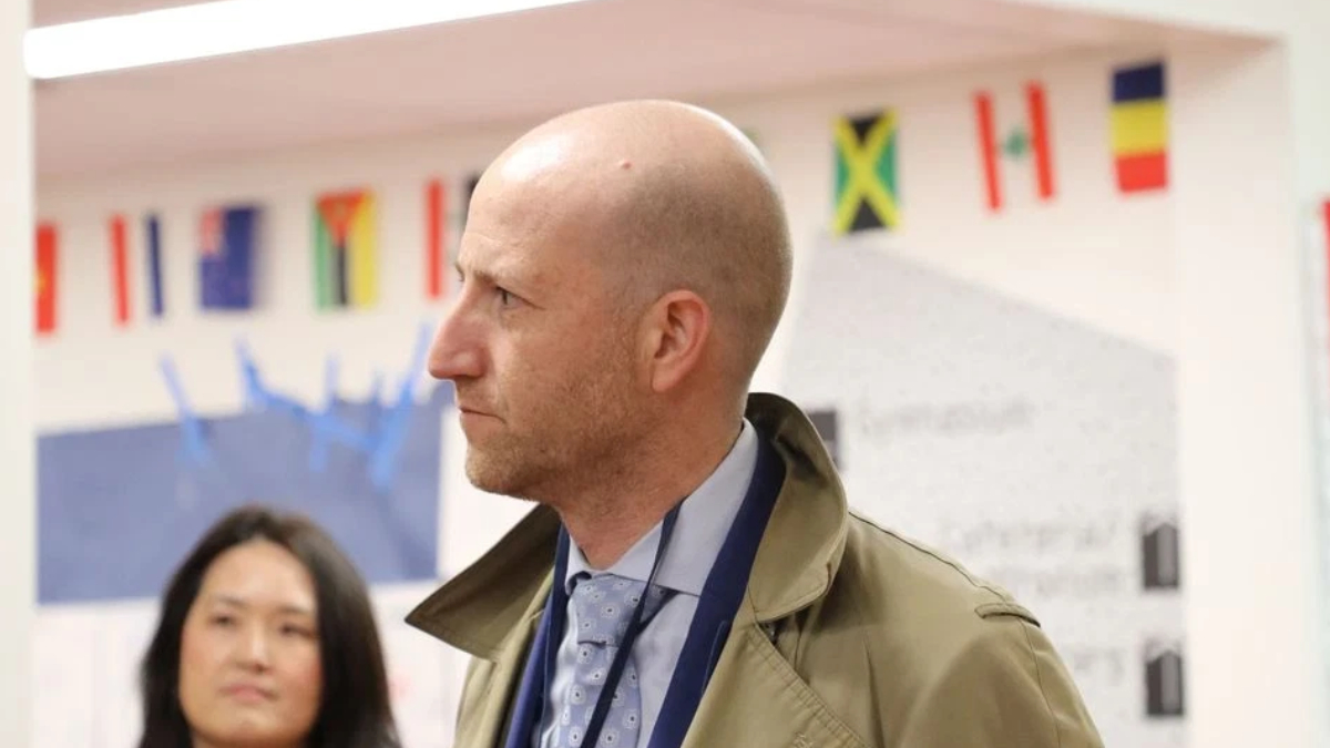 Side profile of a bald man in a blue shirt and tie wearing a tan trench coat at an indoor event, with colorful flags in the background.