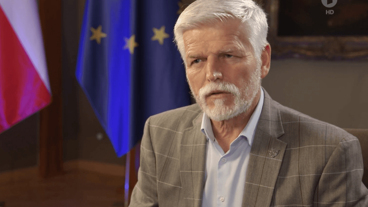 Older man with white hair and beard in a gray plaid suit, seated indoors with EU and national flags in the background.