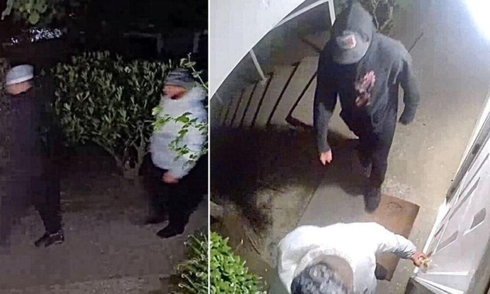 Two masked men standing outside a house at night; one in a gray beanie and white jacket, the other in dark clothing.
