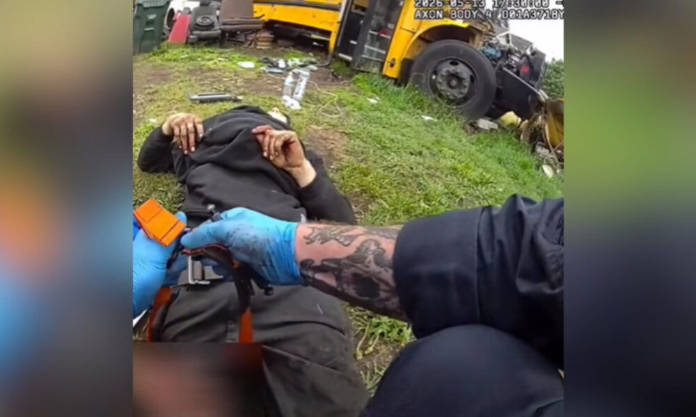 Emergency responder with blue gloves tending to an injured person lying on grass near a yellow bus wreck and scattered debris.