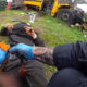 Emergency responder with blue gloves tending to an injured person lying on grass near a yellow bus wreck and scattered debris.
