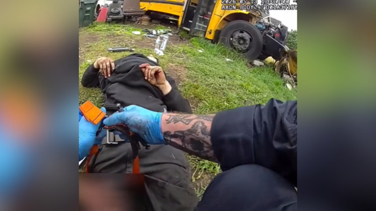 Emergency responder with blue gloves tending to an injured person lying on grass near a yellow bus wreck and scattered debris.