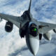 Front-on view of a sleek military fighter jet in mid-flight, nose toward the camera, flying above cloudy skies.