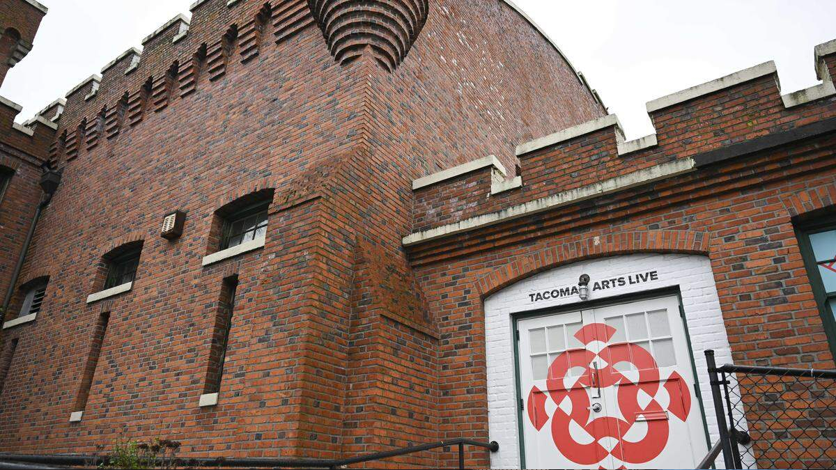 Brick exterior of a historic-style building with battlements and a white door labeled 'Tacoma Arts Live' featuring a red geometric mural on the door surface.