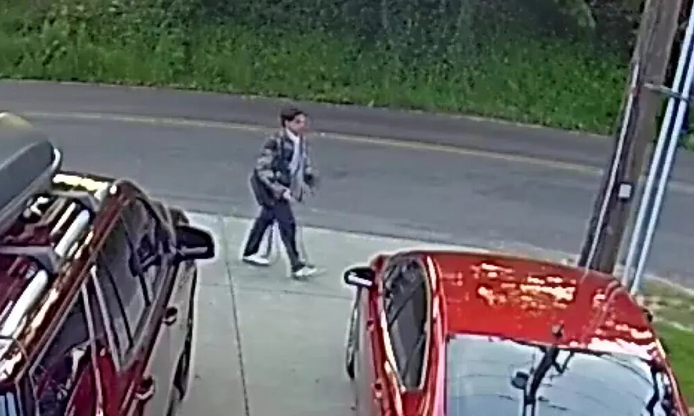 Man in a gray jacket walking on the sidewalk beside parked cars in a lot.
