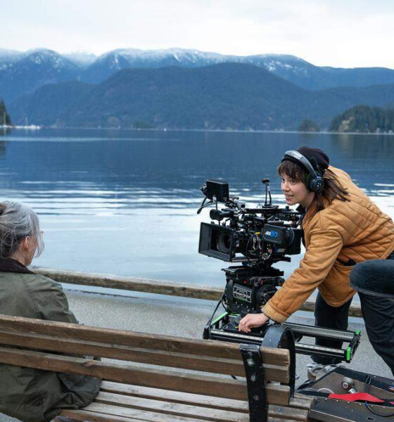 Film crew sets up a large camera rig on a bench beside a calm lake, filming a seated woman with mountains in the distance.