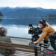 Film crew sets up a large camera rig on a bench beside a calm lake, filming a seated woman with mountains in the distance.