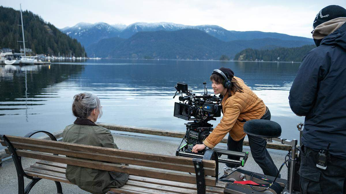 Film crew sets up a large camera rig on a bench beside a calm lake, filming a seated woman with mountains in the distance.