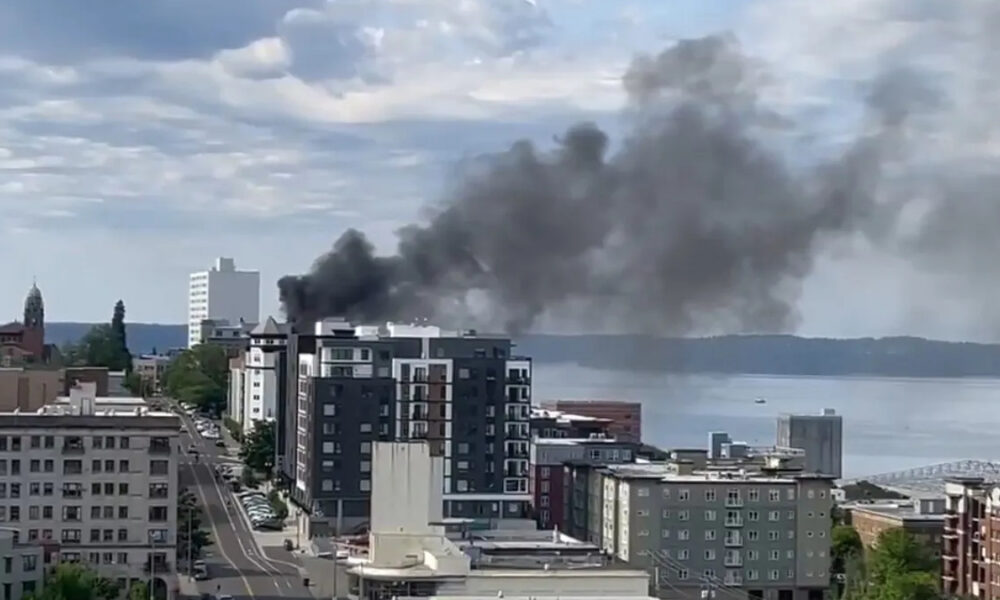 Smoke plume rising from a mid-rise building in a downtown waterfront district, indicating a fire nearby.