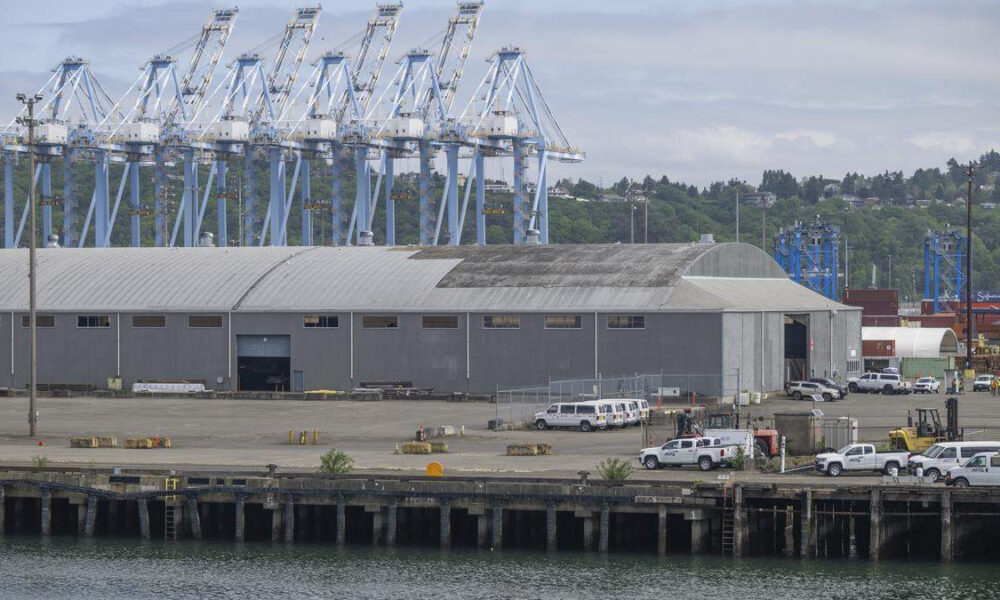 Large gray warehouse by the water with a row of blue gantry cranes in the background and parked trucks along the dock.