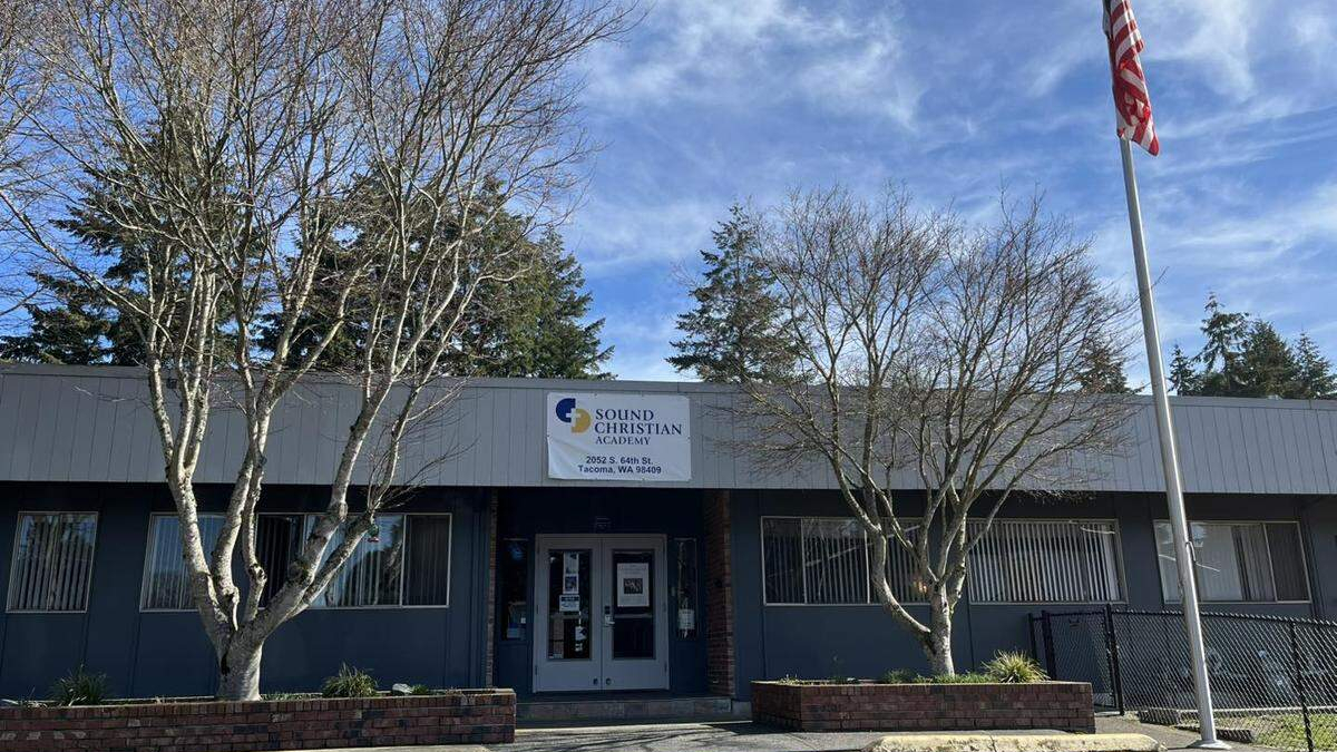 Front view of Sound Christian Academy building with a banner above the entrance, two leafless trees flanking the doors, and an American flag on a tall pole to the right.