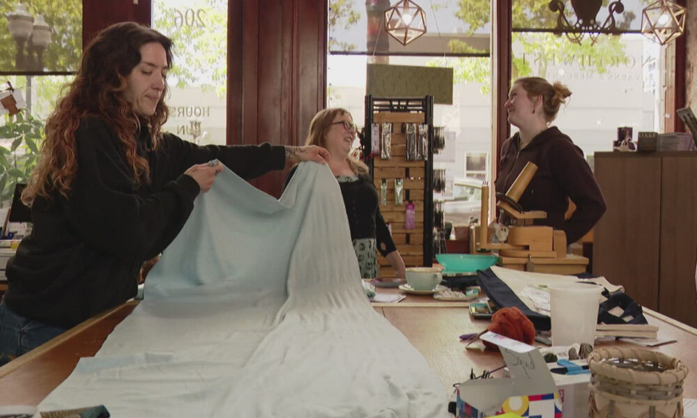 Women in a craft shop unfold a large light-blue fabric on a table, preparing a sewing project while others chat nearby.