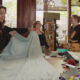 Women in a craft shop unfold a large light-blue fabric on a table, preparing a sewing project while others chat nearby.