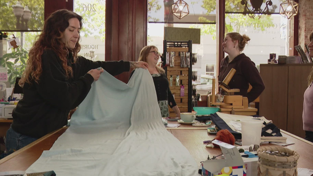 Women in a craft shop unfold a large light-blue fabric on a table, preparing a sewing project while others chat nearby.