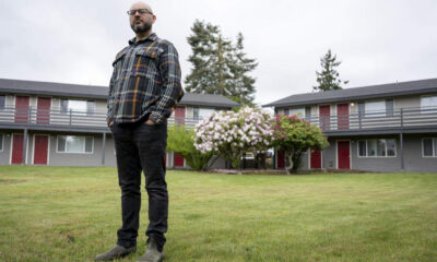 Man with a beard and glasses in a plaid shirt stands on a grassy lawn in front of a two-story apartment building with red doors.