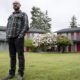 Man with a beard and glasses in a plaid shirt stands on a grassy lawn in front of a two-story apartment building with red doors.