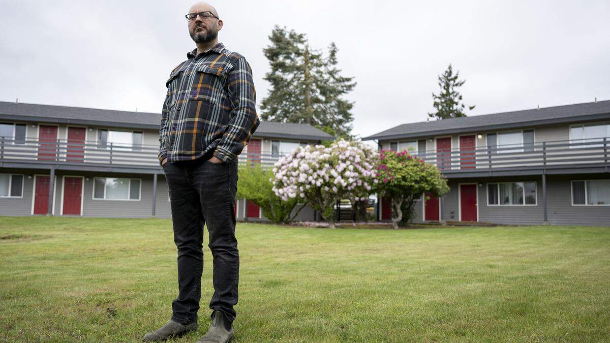 Man with a beard and glasses in a plaid shirt stands on a grassy lawn in front of a two-story apartment building with red doors.