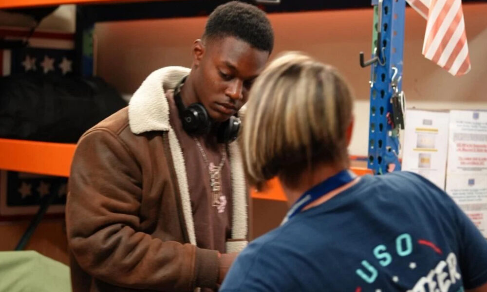 Young man in a brown shearling jacket with headphones looks down at a task as a staff member in a blue USO shirt assists him in a workshop nearby.