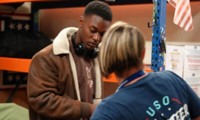 Young man in a brown shearling jacket with headphones looks down at a task as a staff member in a blue USO shirt assists him in a workshop nearby.