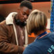 Young man in a brown shearling jacket with headphones looks down at a task as a staff member in a blue USO shirt assists him in a workshop nearby.