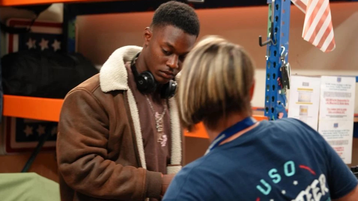 Young man in a brown shearling jacket with headphones looks down at a task as a staff member in a blue USO shirt assists him in a workshop nearby.