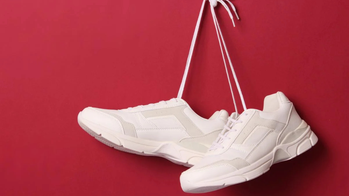 Pair of white sneakers suspended by their laces against a red background.