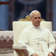 Pope in white robes sitting on an ornate chair in a church setting with an altar behind him
