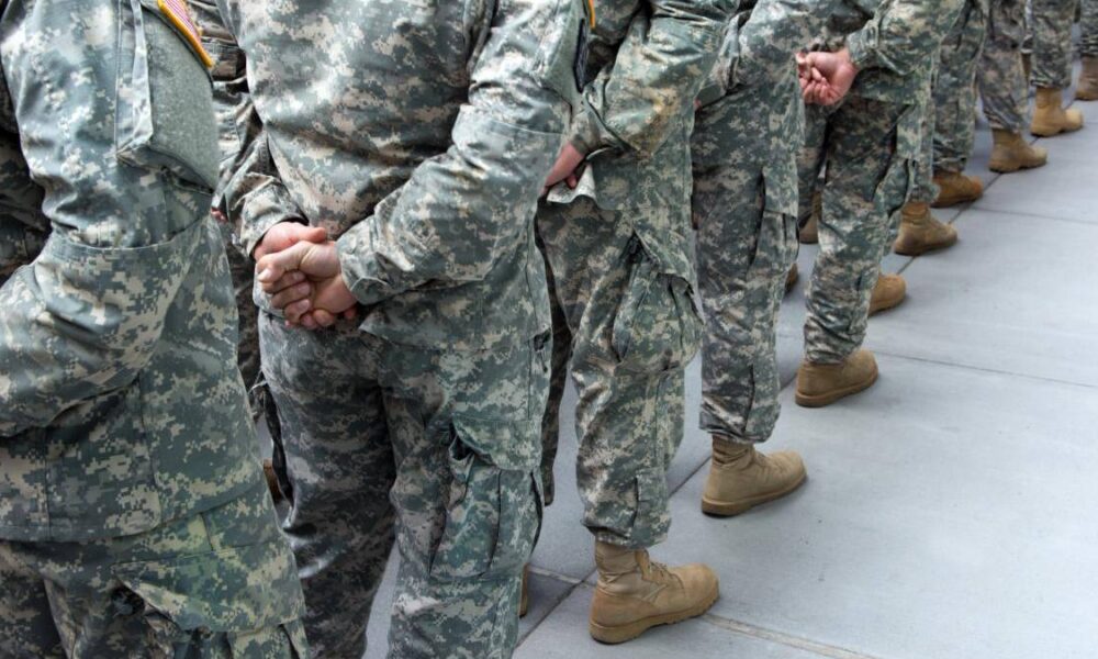 Soldiers in camouflage uniforms standing in a line with hands behind their backs on a concrete surface.