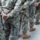 Soldiers in camouflage uniforms standing in a line with hands behind their backs on a concrete surface.