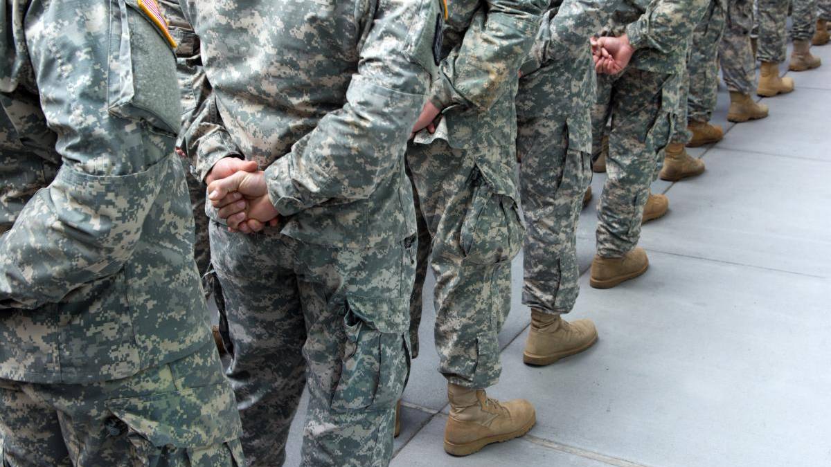 Soldiers in camouflage uniforms standing in a line with hands behind their backs on a concrete surface.