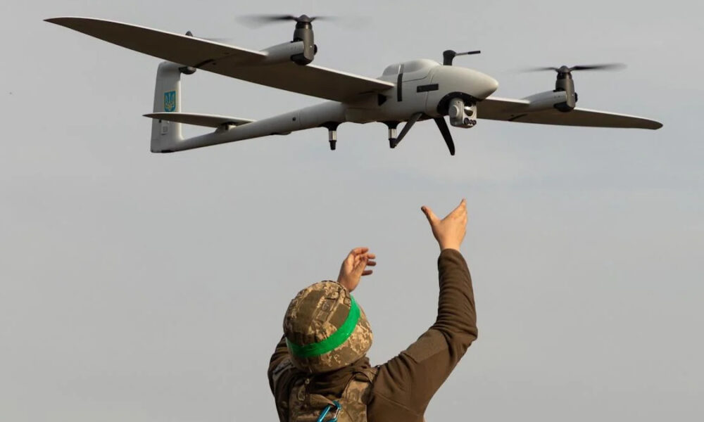 Soldier in camouflage gear and a green headband raises arms toward a quadcopter drone flying overhead.