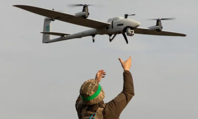 Soldier in camouflage gear and a green headband raises arms toward a quadcopter drone flying overhead.