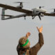 Soldier in camouflage gear and a green headband raises arms toward a quadcopter drone flying overhead.