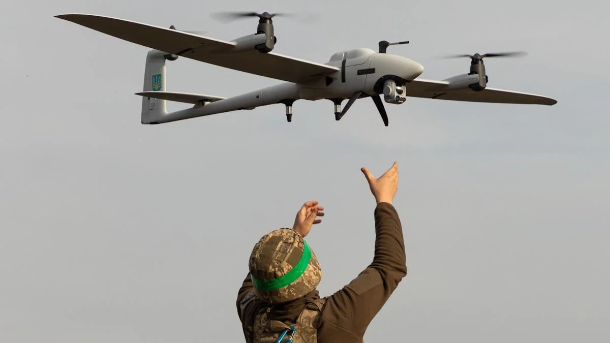 Soldier in camouflage gear and a green headband raises arms toward a quadcopter drone flying overhead.